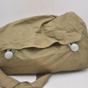 Puede incluir: Un bolso de hombro de lona color caqui con cierre de solapa y correa larga. El bolso tiene dos botones plateados y una correa verde claro. El bolso está hecho de un material duradero y probablemente está diseñado para transportar objetos.