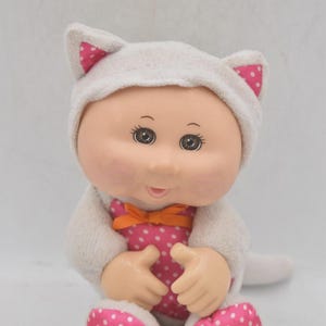 Op de afbeelding: Een pluche pop met een witte kattenorenkap en bijpassende outfit. De outfit van de pop bevat een roze jurk met witte stippen met een oranje strik en roze stippenvoeten. De pop heeft grote ogen en een glimlach.