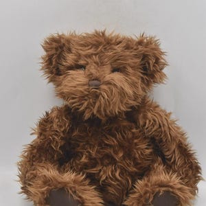 Puede incluir: Un oso de peluche marrón con una apariencia esponjosa y texturizada. El oso tiene una nariz pequeña y redonda y almohadillas oscuras en las patas. El pelaje es de un rico color marrón cálido, y el oso está sentado.