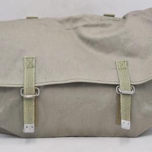 Puede incluir: Bolso mensajero de lona beige con cierre de solapa y dos correas ajustables con hebillas metálicas. El bolso tiene forma rectangular y parece estar hecho de material duradero, adecuado para transportar objetos. El bolso es de color neutro.