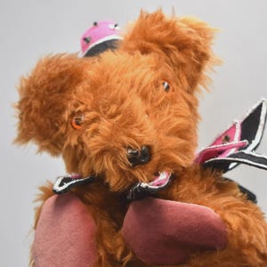 Puede incluir: Un oso de peluche marrón hecho a mano con un sombrero rosa y negro y alas a juego. El oso tiene almohadillas rosas y ojos naranjas. El oso es un artículo único y hecho a mano.