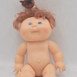 May include: A vintage Cabbage Patch Kid doll with a peach skin tone, blue eyes, and auburn hair styled in a small ponytail. The doll is nude, with a round belly button and rosy cheeks. The doll is sitting down.