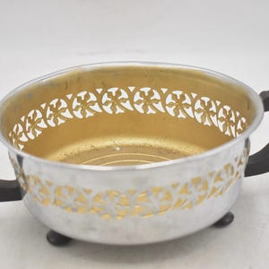Puede incluir: Cuenco plateado con interior dorado y patrón decorativo calado alrededor del borde. El cuenco tiene dos asas negras y descansa sobre tres pequeños pies negros. Una pieza decorativa vintage.
