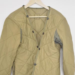 Puede incluir: Chaqueta acolchada de color beige con un patrón de diamantes, cierre de botones y dos bolsillos delanteros. La chaqueta tiene cuello en V y está colgada de una percha de madera. Los puños y el cuello son de color verde oscuro.