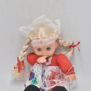 Puede incluir: Muñeca vintage con trenzas rubias atadas con cintas rojas. La muñeca lleva un vestido rojo, un delantal de encaje blanco y un gorro de encaje blanco. La muñeca tiene ojos azules y zapatos negros.