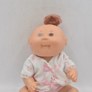 May include: A vintage baby doll with a peach-colored complexion, brown hair in a topknot, and large, expressive eyes. The doll wears a white floral-print jacket with pink bows and matching pink underwear. The doll is sitting down.