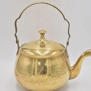 Puede incluir: Una tetera de latón vintage con un diseño floral grabado. La tetera tiene un pico curvo, un asa curva y una tapa con un pequeño pomo. La tetera es de color dorado brillante y probablemente se utiliza para preparar té.