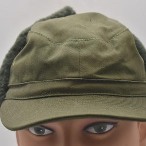 Könnte beinhalten: Olivgrüner Hut im Militärstil mit kurzem Schirm und Ohrenklappen. Der Hut hat eine strukturierte Krone und ein Band um den Ansatz. Die Ohrenklappen sind in einem dunkleren Grünton gehalten und haben eine flauschige Textur.