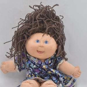 Peut inclure: Une poupée vintage avec un visage doux et rond, des yeux bleus et des cheveux bruns en fil. La poupée porte une robe bleue à motifs floraux avec une bordure blanche. La poupée est assise, les jambes tendues.