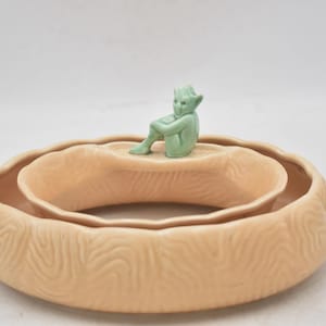 May include: A light peach ceramic planter with a wood-grain texture. A small, seated, green figurine sits on the inner rim. The planter has a layered, circular design. The measurements are in centimetres.
