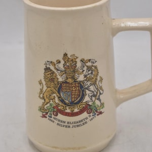 May include: Cream-colored ceramic mug with a handle, featuring a detailed emblem with a crown, lions, and a unicorn. The mug commemorates the Queen Elizabeth II Silver Jubilee, 1952-1977.