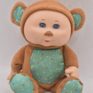 May include: A vintage doll with a brown bear costume. The doll has a light skin tone, blue eyes, and a small brown nose. The bear costume is brown with a teal belly and foot pads decorated with yellow and orange polka dots.