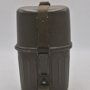 May include: An olive green metal container with a cylindrical top and a ridged lower section. A matching green canvas strap with a metal buckle secures the lid. The container is designed for storage and transport.