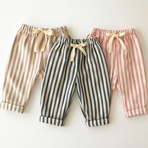 May include: Three pairs of striped baby pants in neutral colors. The pants are in shades of tan, navy and white, and pink and white. Each pair has a cream-colored bow at the waist and cuffed hems.