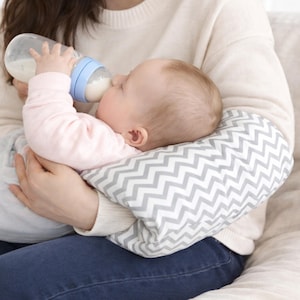 May include: A nursing pillow featuring a white and gray chevron pattern, providing support for a baby who is drinking from a clear plastic bottle with a light blue cap. The pillow is shaped to fit around the arm for comfortable positioning.