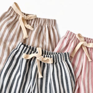 Puede incluir: Primer plano de tres pares de pantalones cortos a rayas en varios colores: marr&oacute;n y blanco, azul marino y blanco y rosa y blanco. Cada par tiene una cintura con cord&oacute;n y un lazo color crema. Los pantalones cortos son de tela ligera.