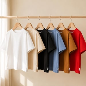 Op de afbeelding: Een rij van zes t-shirts met korte mouwen en een ronde hals die aan houten kleerhangers hangen. De shirts worden getoond in een reeks kleuren: wit, beige, zwart, stoffig blauw, mosterdgeel en rood. Elk shirt heeft een klein wit label.