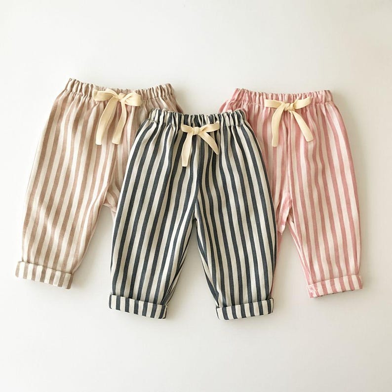 May include: Three pairs of striped baby pants in neutral colors. The pants are in shades of tan, navy and white, and pink and white. Each pair has a cream-colored bow at the waist and cuffed hems.