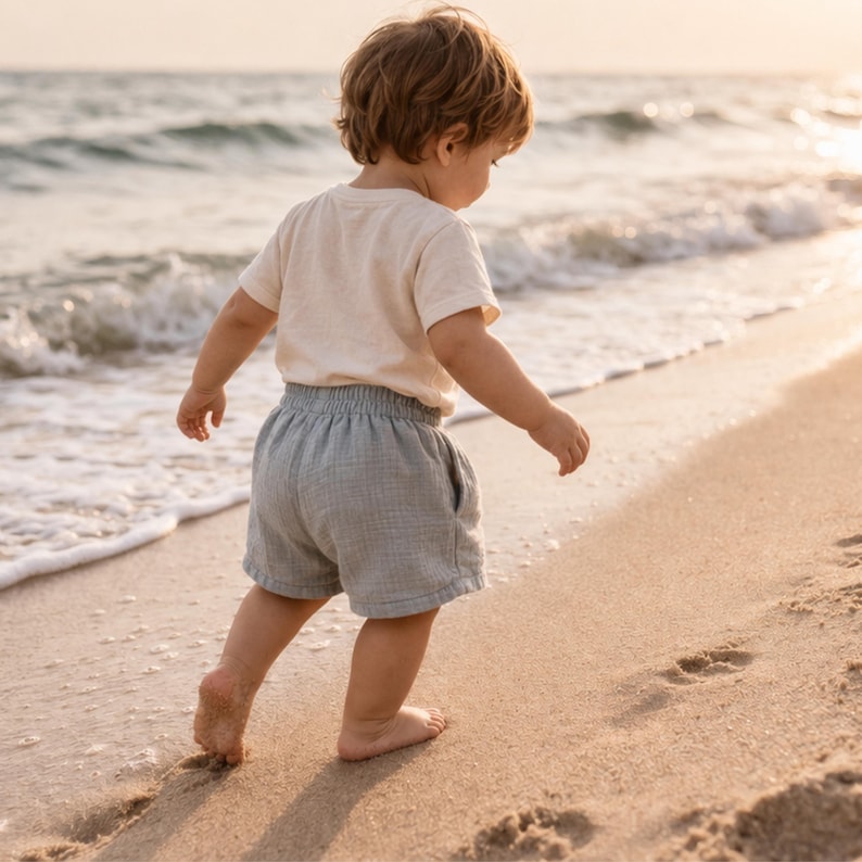 May include: A young child wearing a cream-colored t-shirt and light blue shorts walks barefoot on a sandy beach. The child is facing away from the camera, with the ocean waves in the background. The sun is setting, casting a warm glow.