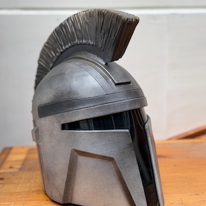 May include: A silver and black helmet with a prominent crest. The helmet has a metallic finish and angular design, reminiscent of a Spartan or Mandalorian helmet. The crest is a dark gray color.