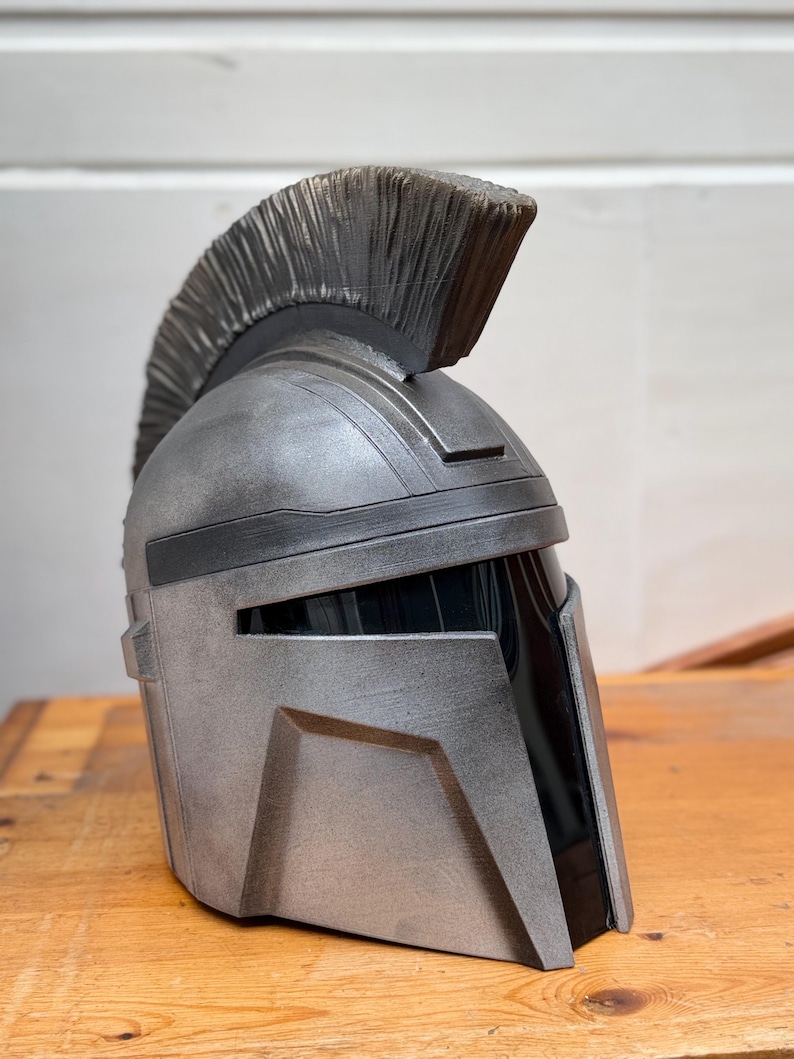 May include: A silver and black helmet with a prominent crest. The helmet has a metallic finish and angular design, reminiscent of a Spartan or Mandalorian helmet. The crest is a dark gray color.