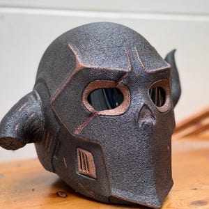 May include: A textured black helmet with copper-coloured accents and two curved horns. The helmet has eye openings and a mouth area, suggesting a skull-like design. The product is likely for cosplay or costume use.