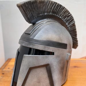 May include: A silver and black helmet with a textured finish and a prominent crest. The helmet has a visor and angular design, reminiscent of a warrior's headgear. The helmet is displayed on a wooden surface.