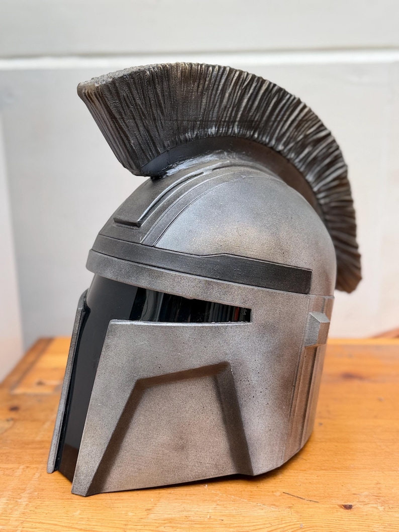 May include: A silver and black helmet with a textured finish and a prominent crest. The helmet has a visor and angular design, reminiscent of a warrior's headgear. The helmet is displayed on a wooden surface.