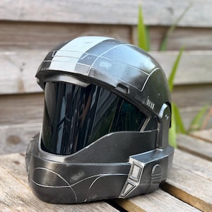 May include: A dark grey helmet with a black visor, set on a wooden surface. The helmet has a metallic sheen and a white stripe across the top. The design features angular lines and a futuristic aesthetic, suggesting a science fiction or cosplay theme.