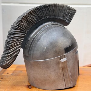 May include: A silver-colored helmet with a textured, dark gray crest. The helmet has a metallic finish and a rectangular visor. The crest is made of numerous, closely-packed, vertical, textured elements.