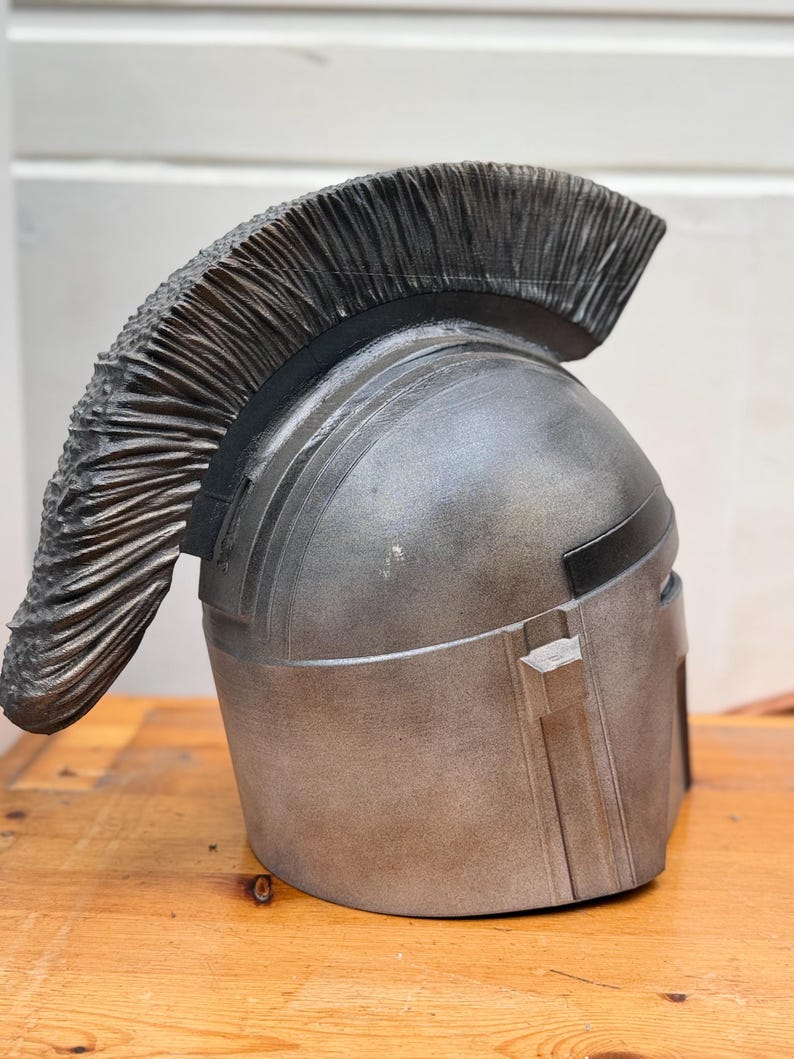 May include: A silver-colored helmet with a textured, dark gray crest. The helmet has a metallic finish and a rectangular visor. The crest is made of numerous, closely-packed, vertical, textured elements.