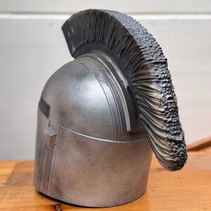 May include: A silver-colored helmet with a dark, textured crest. The helmet has a rectangular detail on the side and is set against a wooden surface.
