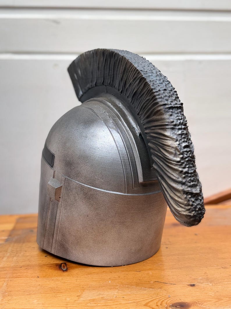 May include: A silver-colored helmet with a dark, textured crest. The helmet has a rectangular detail on the side and is set against a wooden surface.
