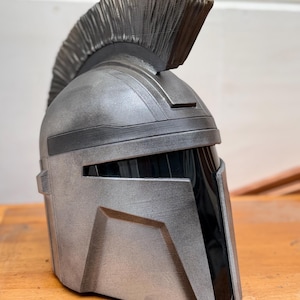 May include: A silver and black helmet with a prominent crest. The helmet has a metallic finish and angular design, with a dark visor area. The crest is a textured, dark gray color. The helmet is likely a costume or collectible item.