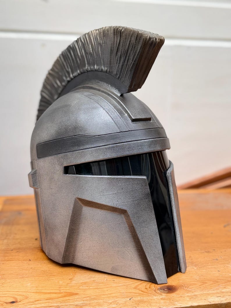 May include: A silver and black helmet with a prominent crest. The helmet has a metallic finish and angular design, with a dark visor area. The crest is a textured, dark gray color. The helmet is likely a costume or collectible item.