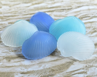 May include: Five frosted glass seashell decorations in shades of blue and white. The shells have a ribbed texture and are arranged on a textured, neutral-toned surface. The colors range from light blue to a deep blue.