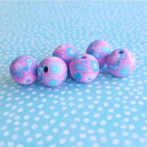 May include: Six colorful beads with a pink, blue, and white abstract pattern. The beads are made of polymer clay and have a hole in the center for stringing.