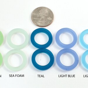 K&ouml;nnte beinhalten: Acht Glasringe in verschiedenen Gr&uuml;nt&ouml;nen, Blau- und Lilat&ouml;nen. Die Ringe sind von links nach rechts in einer Reihe angeordnet, wobei die Farben allm&auml;hlich von Gr&uuml;n nach Lila &uuml;bergehen. Die Ringe sind alle gleich gro&szlig; und gleich geformt und bestehen alle aus klarem Glas. Die Farben sind unter den Ringen beschriftet: Flaschengr&uuml;n, Hellgr&uuml;n, Meeresschaum, Teal, Hellblau, Hellblau, Lavendel.
