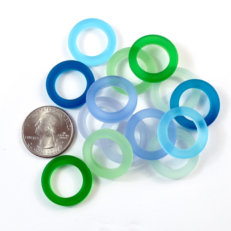 K&ouml;nnte beinhalten: Eine Sammlung von zehn Glasringen in verschiedenen Blaut&ouml;nen und Gr&uuml;nt&ouml;nen. Die Ringe sind alle ungef&auml;hr gleich gro&szlig; und liegen lose &uuml;bereinander.