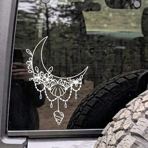 May include: A white vinyl decal applied to a vehicle window, featuring a crescent moon shape adorned with floral elements, hanging beaded strands, and a central seashell motif, with a single spiral shell dangling from the bottom centre.