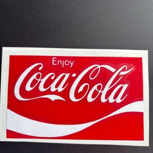 May include: A rectangular sticker featuring the Coca-Cola logo in white against a red background. The word "Enjoy" is above the logo. The sticker has a white border.