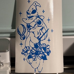 Pode incluir: Uma ilustração azul e branca de um sapo usando um chapéu de mago com estrelas no fundo. O sapo está segurando uma varinha e está rodeado de flores.