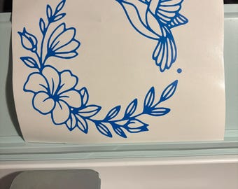 Puede incluir: Un diseño azul de colibrí y corona floral sobre fondo blanco. El colibrí está en vuelo, rodeado de hojas, flores y puntos. El diseño es probablemente una calcomanía o pegatina, adecuado para varios proyectos de manualidades.