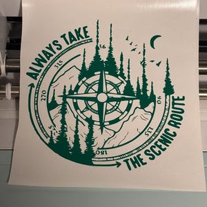 以下が含まれることがあります： コンパスのデザイン、山、木、そして「ALWAYS TAKE THE SCENIC ROUTE」というテキストが特徴の緑色のビニールステッカー。デザインには三日月と鳥が含まれています。ステッカーは白い背景にあります。