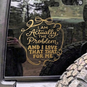 Peut inclure: Un autocollant en vinyle doré appliqué sur une vitre de véhicule, présentant une bordure décorative avec un nœud en haut et le texte : I AM Actually THE Problem AND I LOVE THAT THAT FOR ME, écrit dans une police stylisée et cursive.