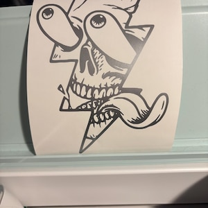 May include: A black and white sticker featuring a stylized skull within a lightning bolt shape. The skull has exaggerated eyes and a long tongue, giving it a cartoonish appearance. The sticker is on a roll, ready for application.