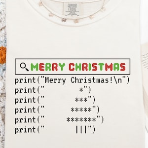 May include: Ivory-colored t-shirt with a graphic that reads "MERRY CHRISTMAS" in a search bar design. Below the text is a computer code-style Christmas tree. A delicate necklace with small butterfly charms is draped near the neckline.