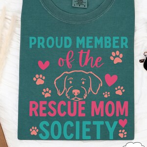 May include: Teal Comfort Colors t-shirt with the text "PROUD MEMBER of the RESCUE MOM SOCIETY" in teal and pink. The design includes a dog illustration, hearts, and paw prints.