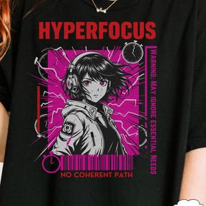May include: Black t-shirt with a graphic featuring an anime-style character wearing headphones, set against a pink and gray background with the text "HYPERFOCUS" and "WARNING: MAY IGNORE ESSENTIAL NEEDS". The design includes clocks and lightning bolts.