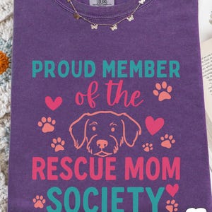 May include: A purple Comfort Colors t-shirt with the text "PROUD MEMBER of the RESCUE MOM SOCIETY" in teal and pink. The design includes paw prints, hearts, and a dog illustration. A gold butterfly necklace is draped on the shirt.